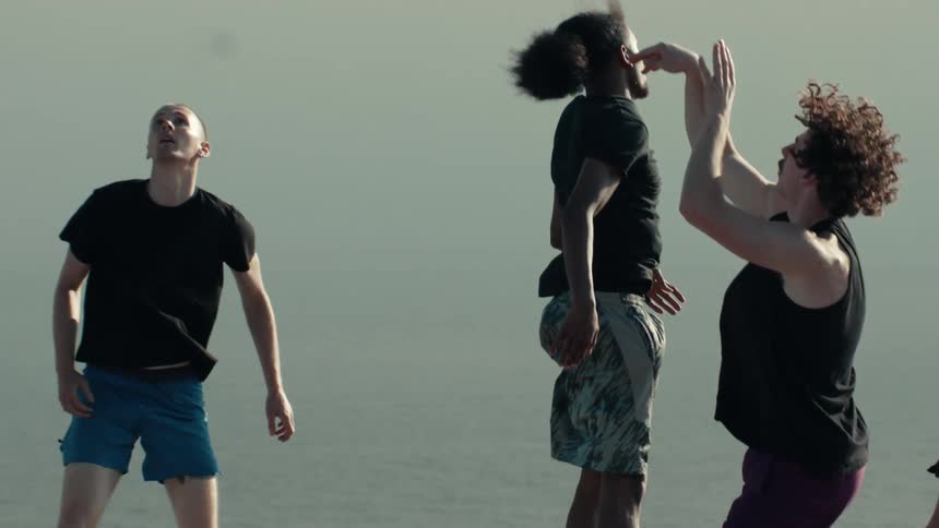 A diverse group of young men play a competitive game of basketball on a seaside court in super slow motion