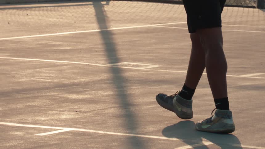 Cinematic slow motion view of a basketball player's feet and a bouncing ball on an outdoor court during golden hour