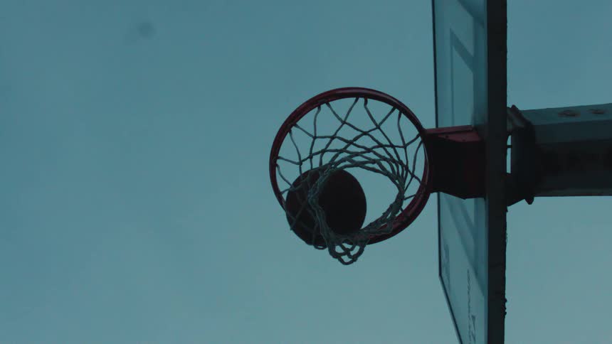 From a low angle, a basketball player dunks a ball in dramatic slow motion against a cool, blue sky