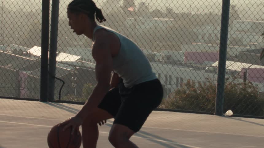 An athletic man dribbles a basketball in slow motion on a fenced-in, outdoor court in the city at golden hour