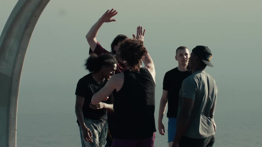 A diverse group of friends laugh and high-five in a celebratory huddle on an outdoor basketball court by the water in slow motion