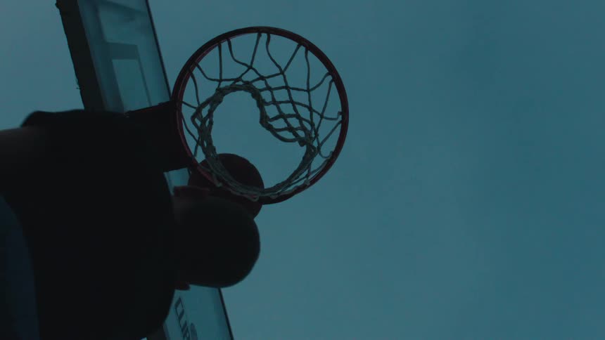 From a low angle, a silhouetted basketball player makes a perfect shot in slow motion against the twilight sky