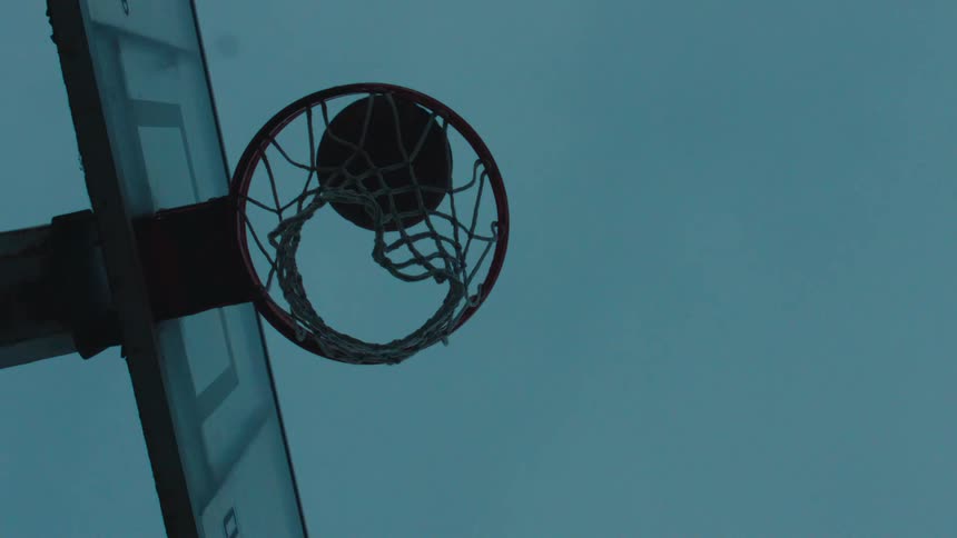 A dramatic slow-motion low-angle shot of a basketball swishing through the net against a cool, dim sky