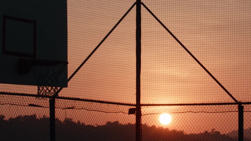 A silhouetted basketball court with a chain-link fence is seen against a beautiful orange sky as the sun rises