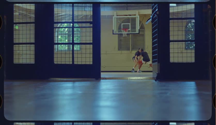 A man and a woman play a competitive game of one-on-one basketball on an indoor court, framed by large industrial doors