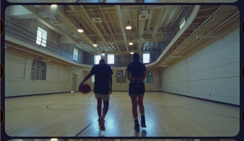 From the shadows into the light, a male and female basketball player walk onto an empty gymnasium court