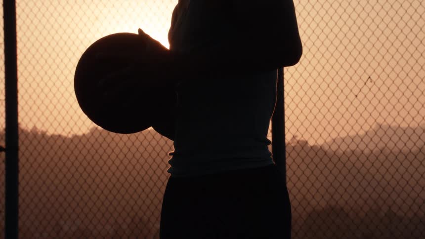 A silhouetted basketball player stands holding a ball against the golden sunset on an outdoor court with a chain-link fence