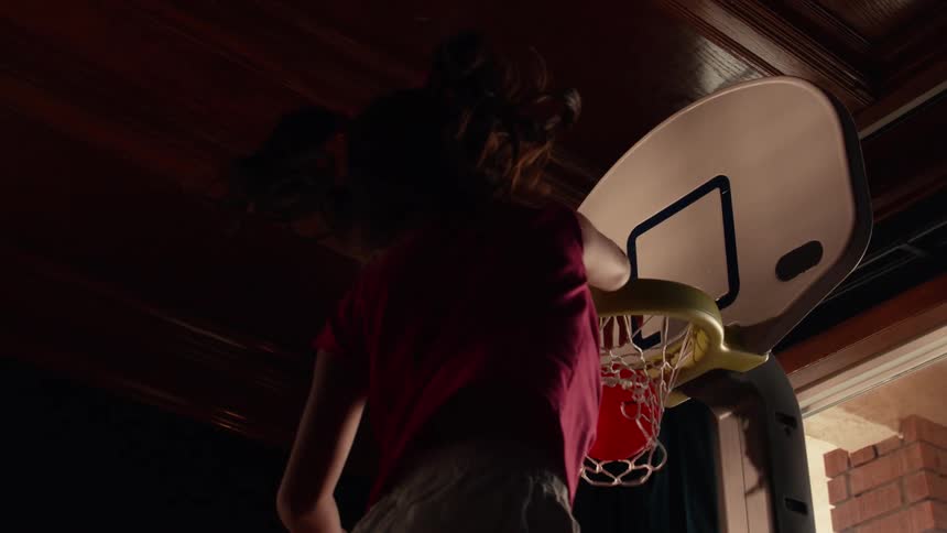 Dramatic slow motion close up from a low angle shows a young girl dunking a basketball into a toy hoop indoors