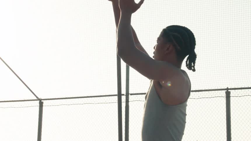 A focused young man shoots a basketball in slow motion on a fenced-in outdoor court against the bright sky