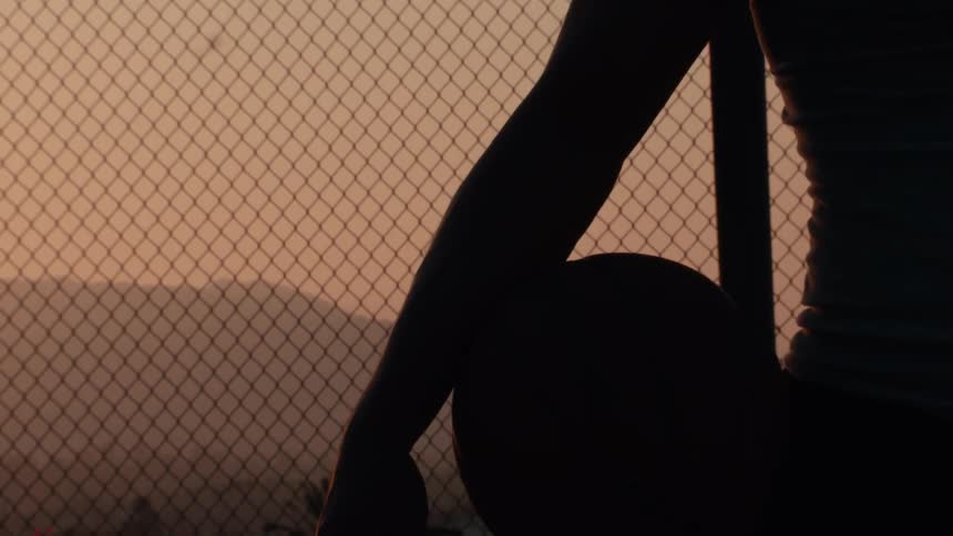 A slow-motion cinematic pull-out shot reveals a silhouetted basketball player holding a ball against a chain-link fence at sunset
