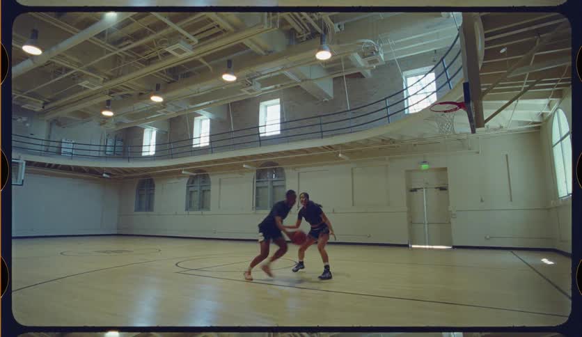 A man skillfully makes a layup in slow motion while playing a one-on-one basketball game against a woman in a vintage gymnasium