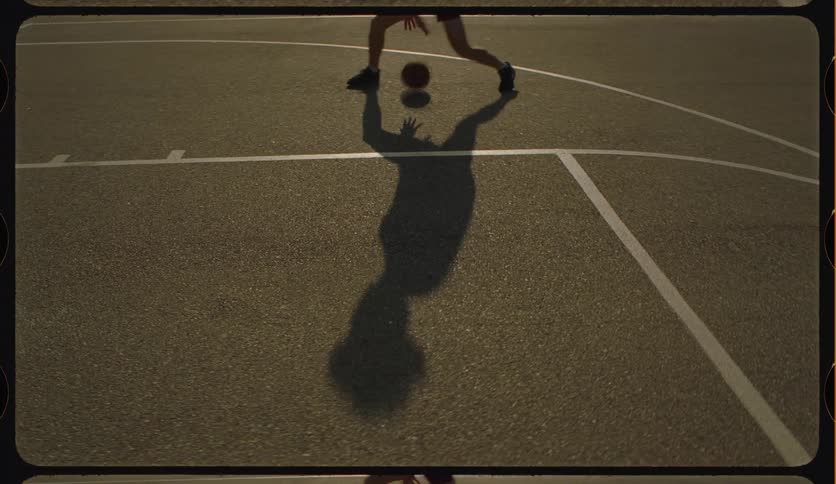 The long shadow of a person dribbles a basketball across an outdoor court in a vintage-style film shot at sunset