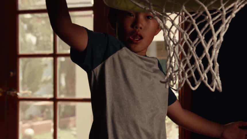 A young Asian boy practices his slam dunk on a small indoor basketball hoop in super slow motion