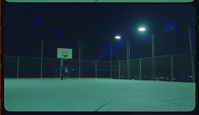 A basketball player practices alone on an outdoor court at night, dribbling towards the hoop and making a slam dunk
