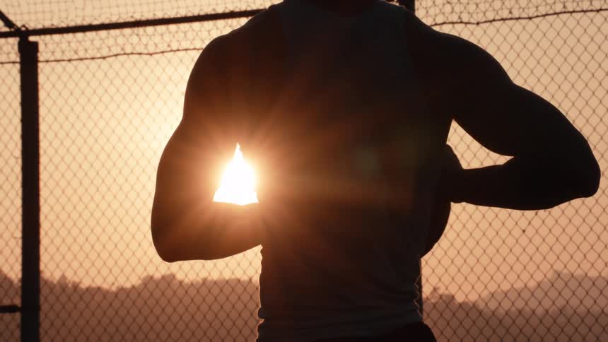 A silhouetted basketball player holds a ball in slow motion, standing before a chain-link fence as the sun sets behind him