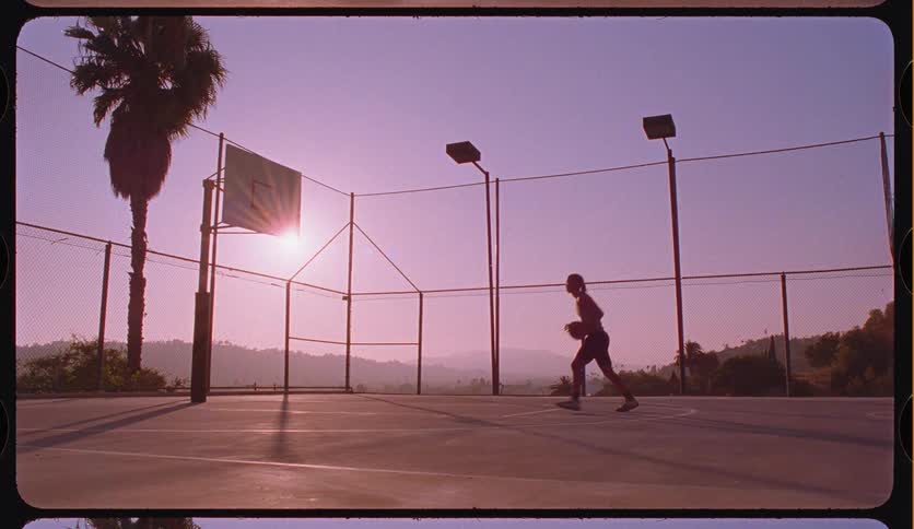 A basketball player shoots hoops in slow motion on a scenic outdoor court, silhouetted against a vibrant purple sunset