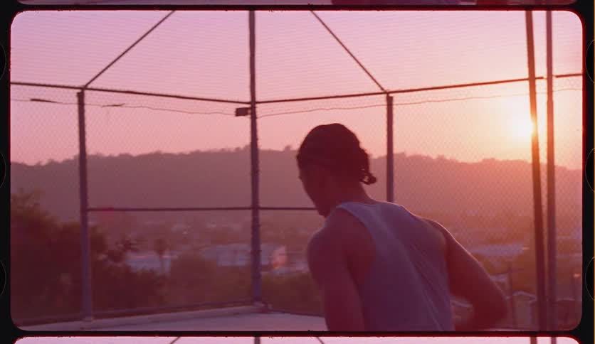 A young man with braided hair shoots a basketball in slow motion on an outdoor court during a beautiful pink sunset