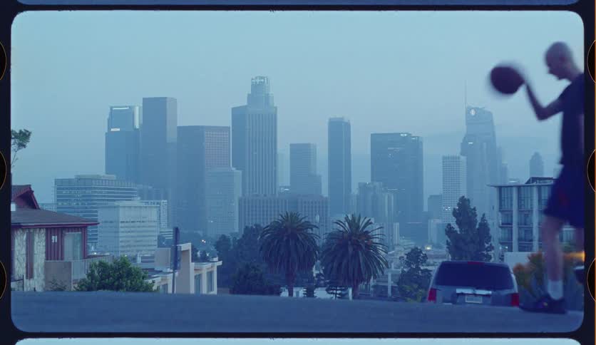 A man dribbles a basketball down a street with the hazy downtown Los Angeles skyline in the background