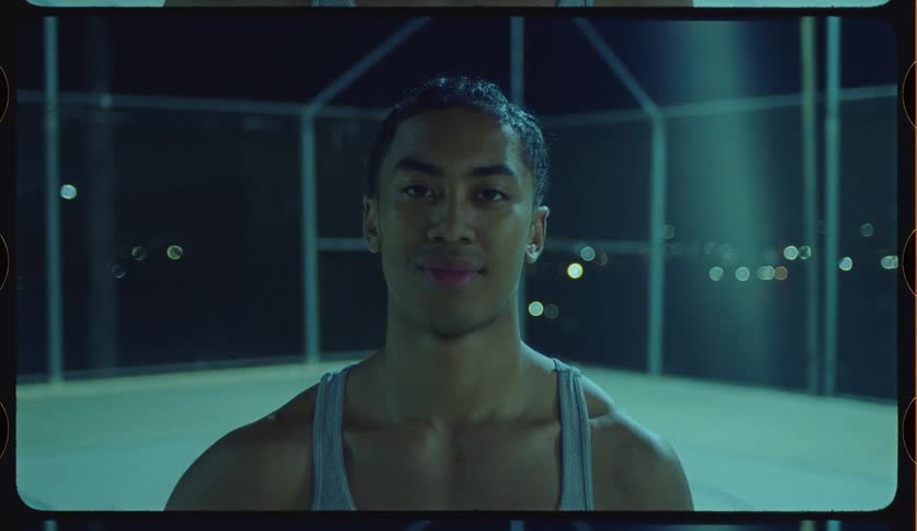 A confident and muscular young athlete looks directly into the camera while standing on an outdoor court at night