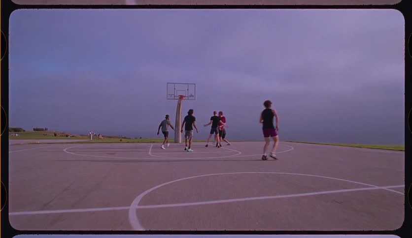 A group of friends play a casual game of pickup basketball on a seaside court in a slow-motion, vintage film-style shot