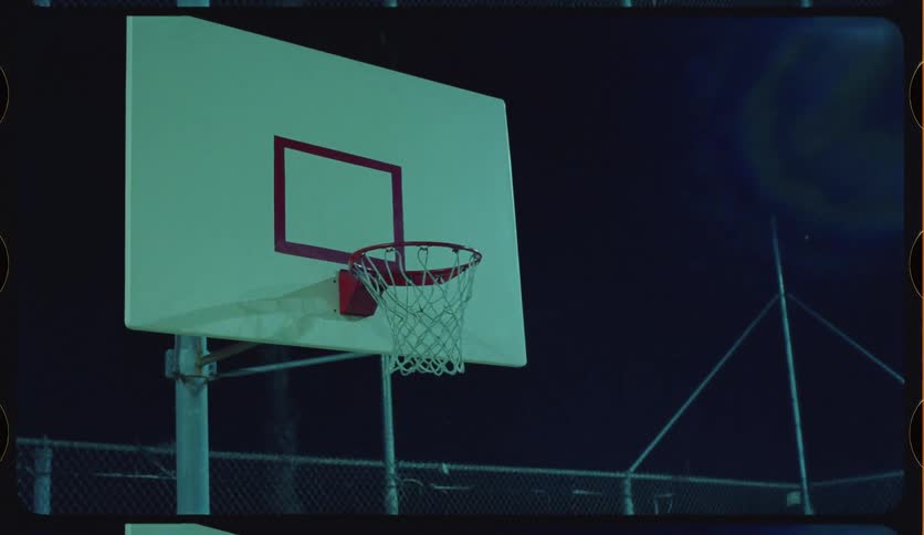 A man dunks a basketball in slow motion on an outdoor court overlooking the city lights at night, shot on vintage film