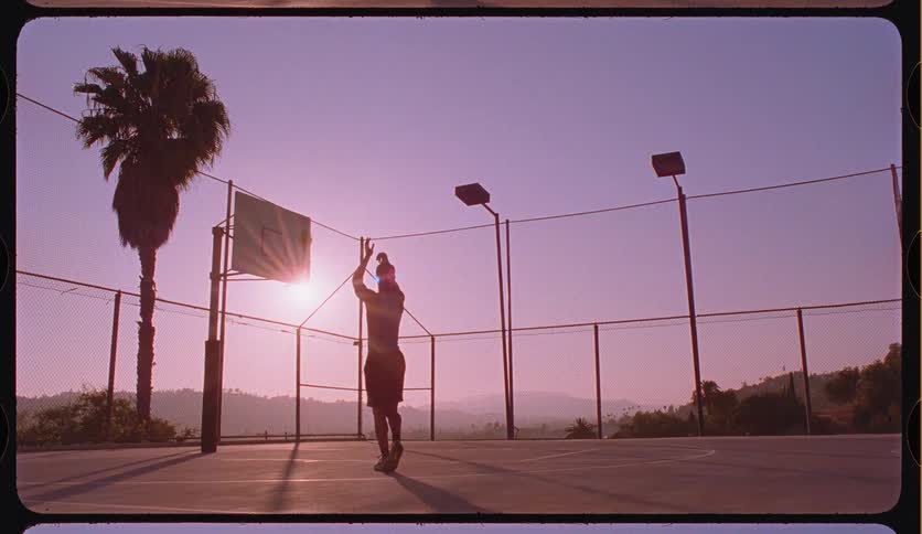 A silhouetted man plays basketball in slow motion on an outdoor court, making a shot against a beautiful purple sunset with a palm tree in the background