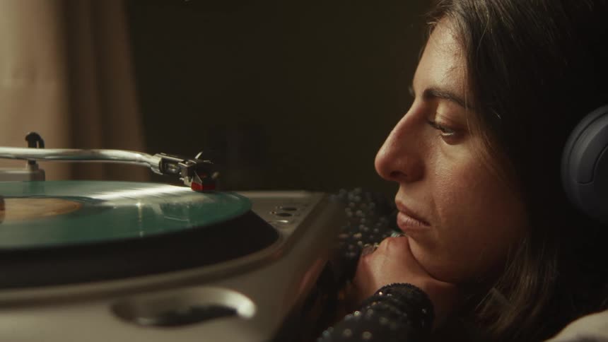 A woman with headphones on rests her head on her hands and sadly listens to music from a spinning vinyl record player