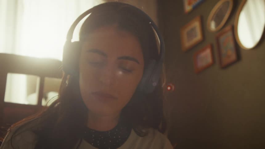 A young woman puts on headphones and closes her eyes to relax while listening to music in a sunlit room