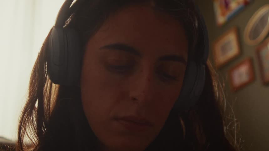An introspective young woman wears headphones and listens with her eyes closed in a warmly lit room
