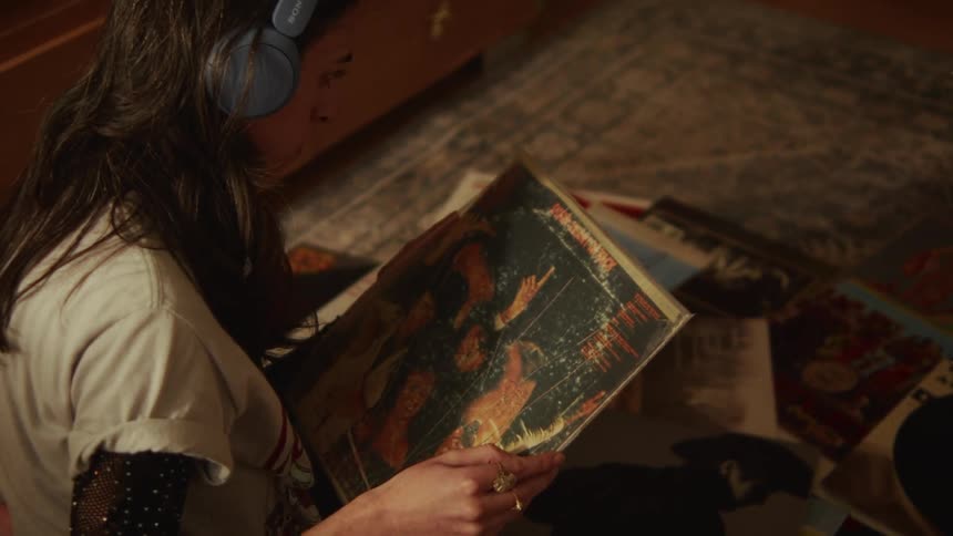 A young woman wearing headphones looks through her vinyl record collection while sitting on the floor in her room