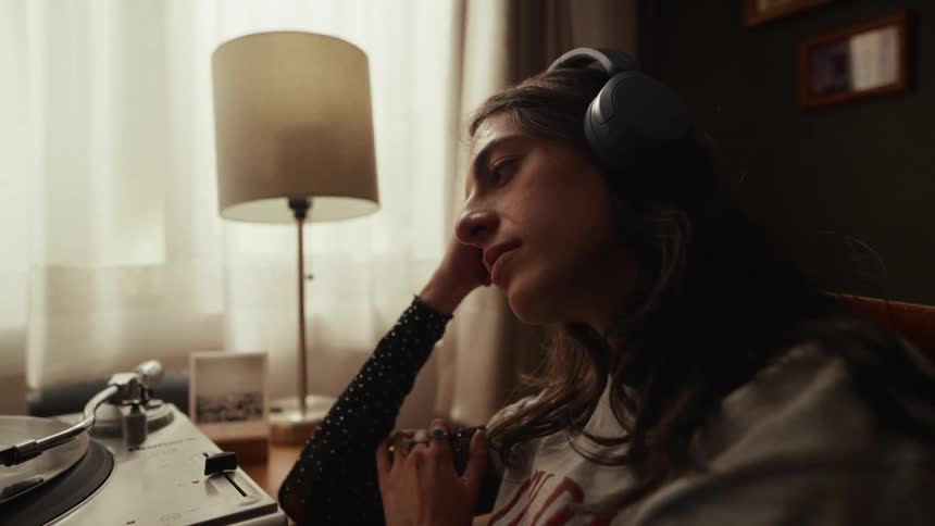 A young woman with headphones on relaxes while listening to a vinyl record spinning on a turntable in a dimly lit room
