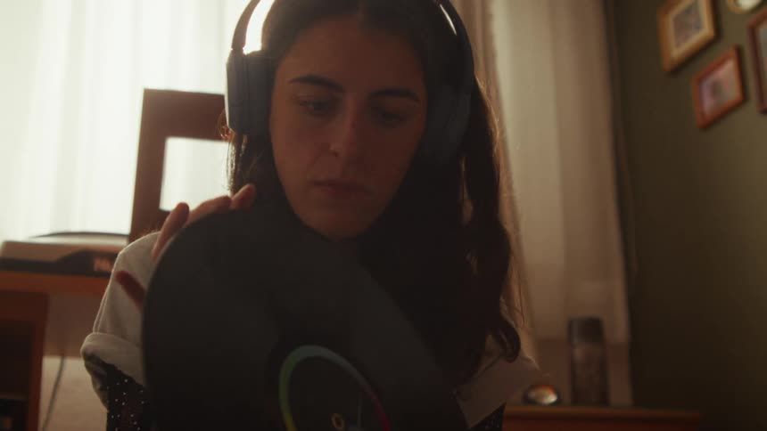 A young woman wearing headphones carefully blows the dust off a vinyl record in a sunlit room