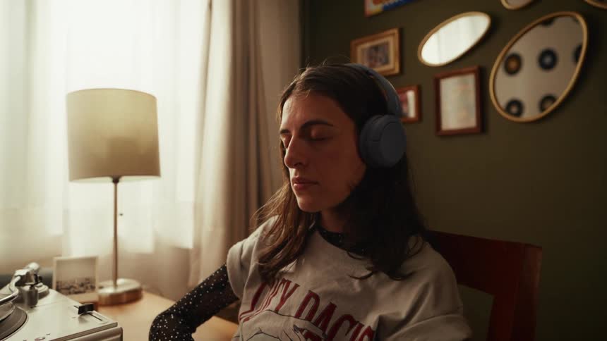 A relaxed young woman with her eyes closed listens to music on headphones from a record player in her cozy room