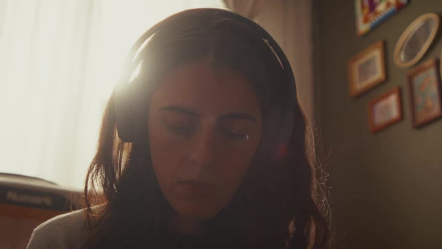 A young woman with headphones closes her eyes, lost in the music she's listening to in a sunlit room