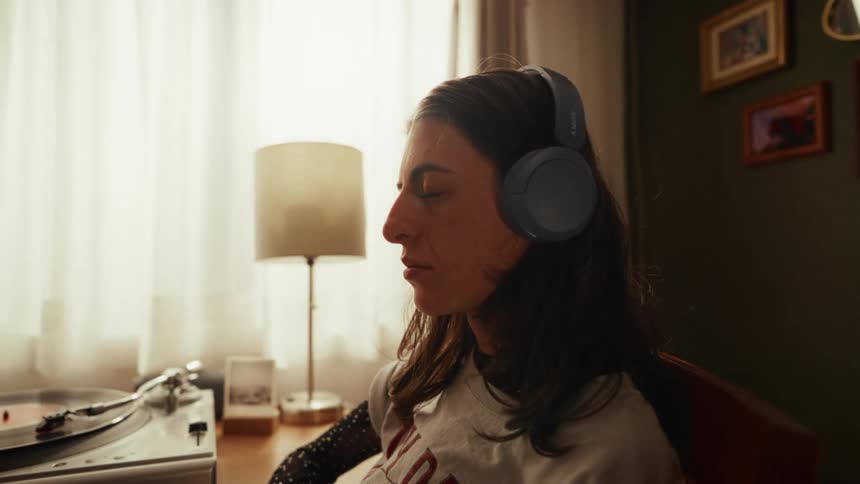 A relaxed woman with her eyes closed peacefully enjoys listening to music from a vinyl record player with headphones on in her room