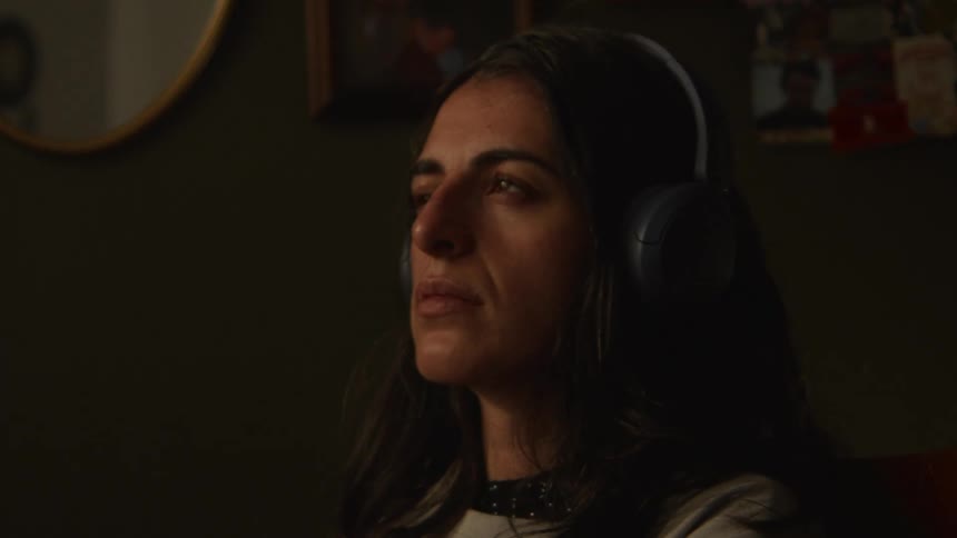 A woman wearing headphones in a dimly lit room opens her eyes after listening intently, revealing a thoughtful expression