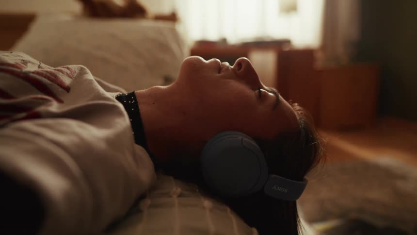 A young person wearing headphones lies on their back in bed, peacefully listening to music in a sunlit room