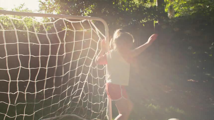 A happy young girl smiles in front of a soccer goal before playfully kicking and throwing the ball in her sunny backyard