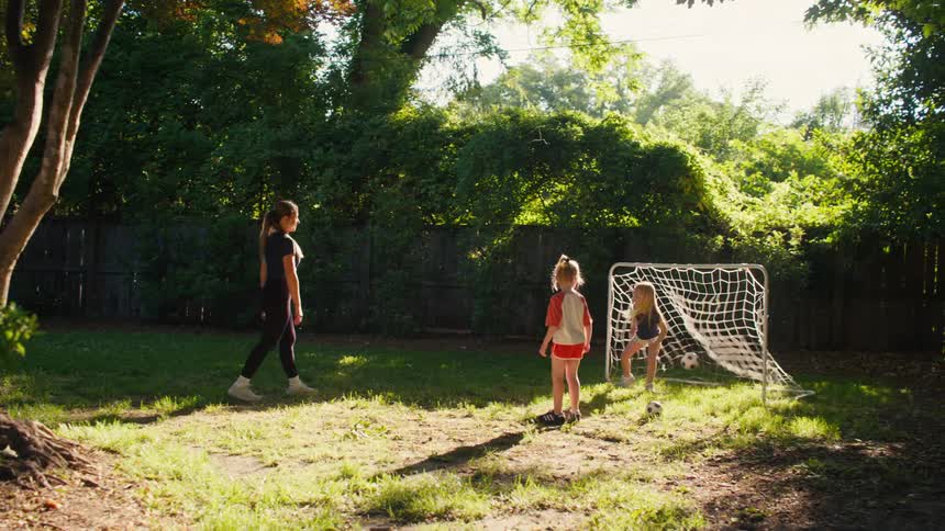 An older sister teaches her two younger siblings how to play soccer in the backyard on a sunny day