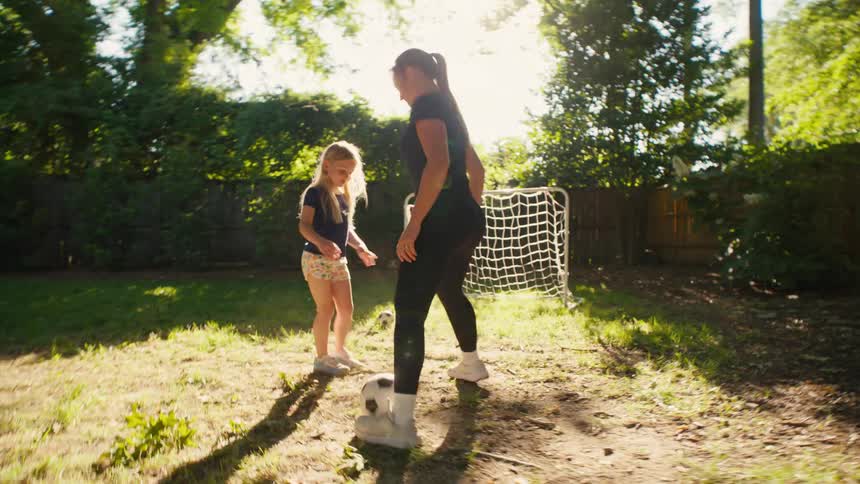 A woman and a young girl practice dribbling a soccer ball together in a sunny backyard