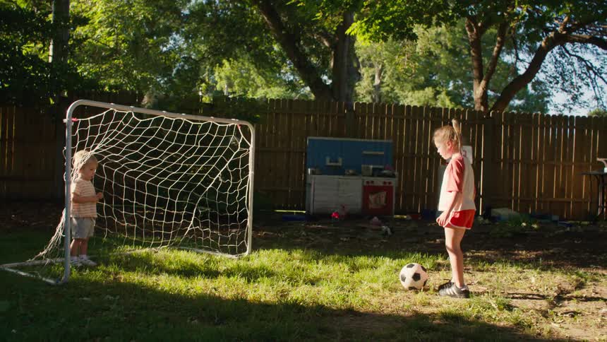 An adorable young girl scores a soccer goal on her toddler brother in the backyard and celebrates her victory