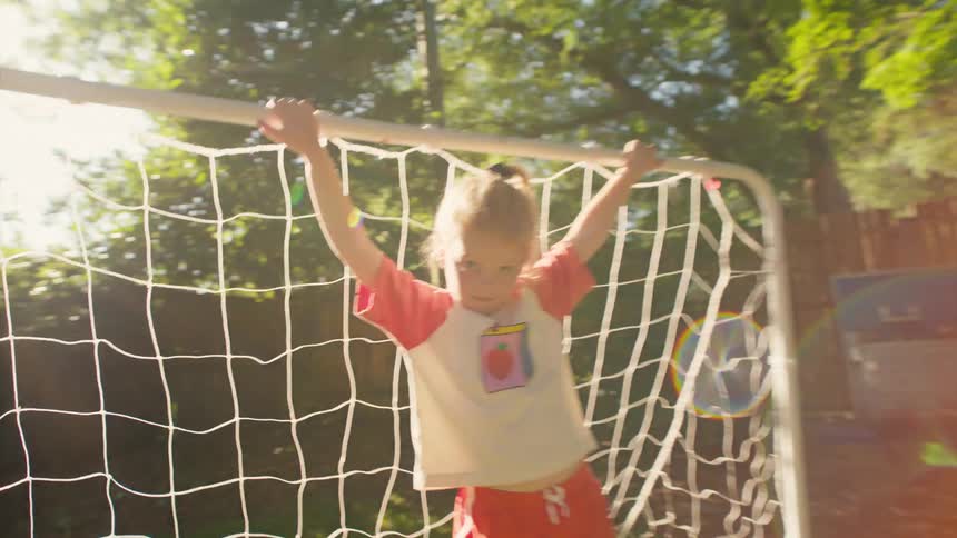 A determined young girl hangs on a soccer goal in her sunny backyard, looking seriously into the camera