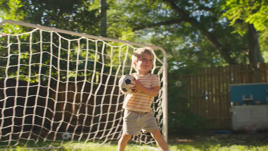 In a sun-drenched backyard, a toddler goalie emerges from the net with his soccer ball and gives it a triumphant throw