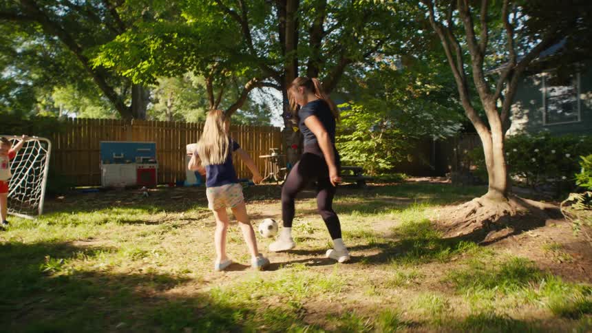 A mother and her two young daughters laugh and play an energetic game of soccer in their sunny backyard