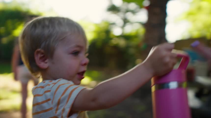 A sad toddler boy gets help with his water bottle from his older sister who then takes a drink while playing in their backyard