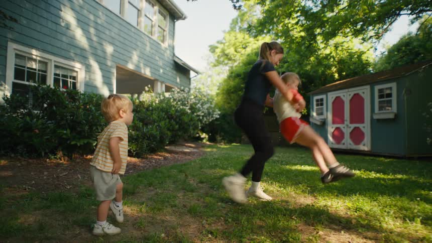 A young mother playfully spins her daughter in circles in their sunny backyard as her toddler son watches, then they play soccer together