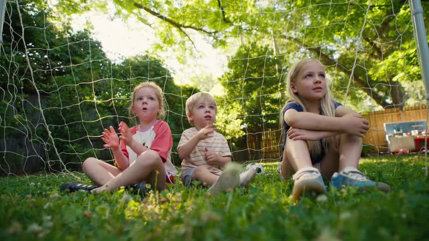 Three young siblings sit inside a soccer goal in their grassy backyard, clapping and looking around with wonder on a sunny summer day