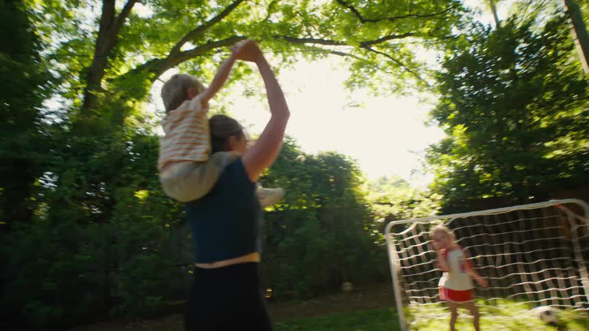 A joyful mother plays soccer with her daughter in their backyard, while carrying her happy toddler son on her shoulders on a sunny day