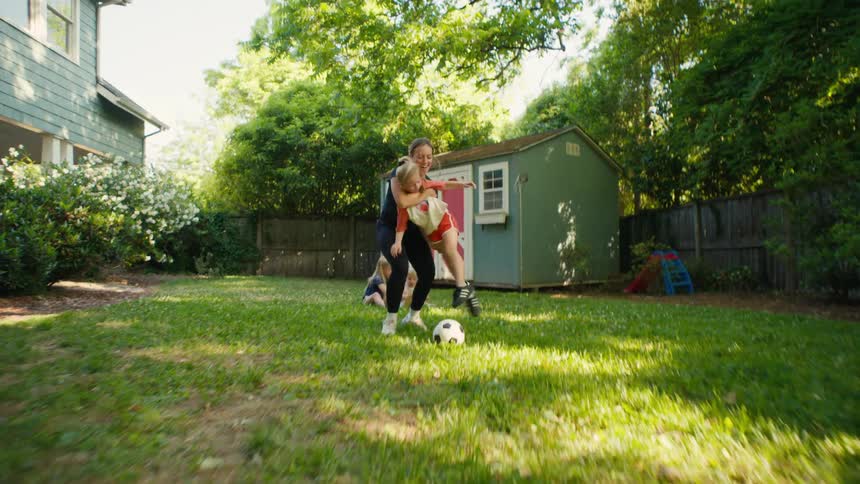 A mother and her young daughter playfully chase a soccer ball in their backyard before the mom picks up her daughter and scores a goal together