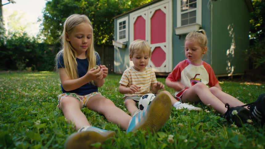 Three adorable young siblings sit on the lush green grass of their backyard, playing with a small soccer ball and making clover chains